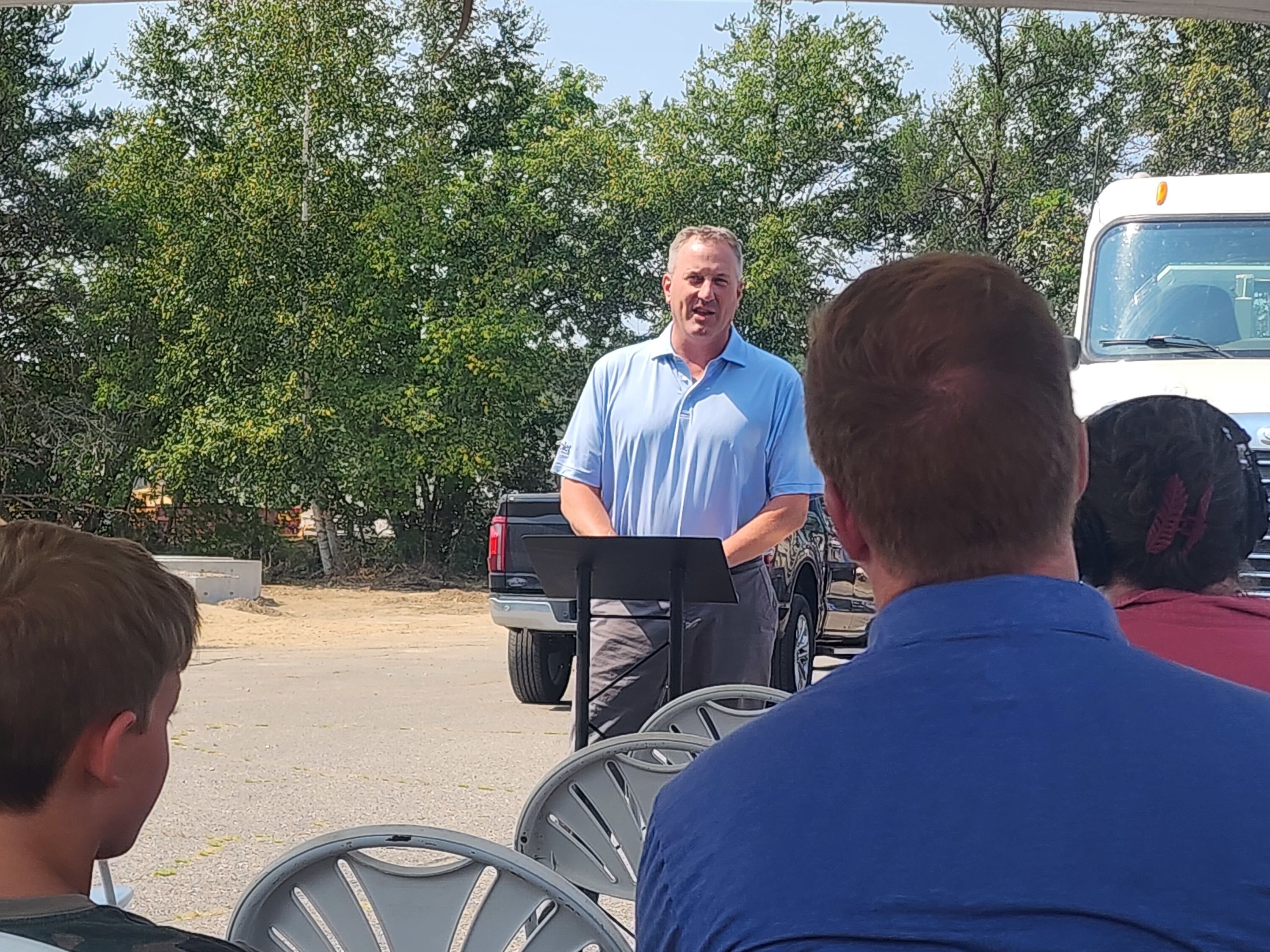 A man in a blue shirt speaks at a podium to an audience seated under a canopy, with vehicles and trees in the background.