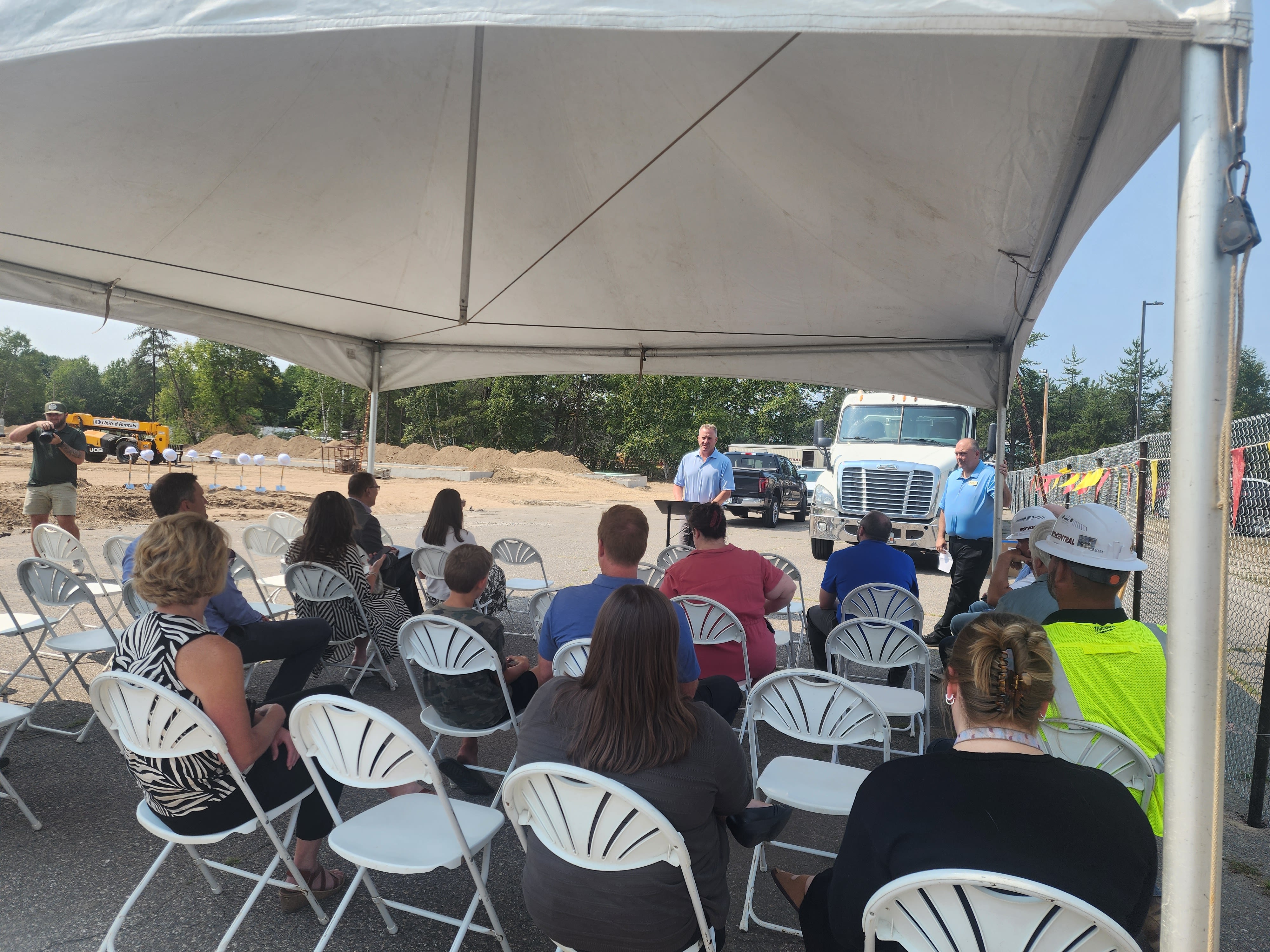 A group of people seated under a large tent, listening to a speaker at an outdoor event site with construction vehicles and wooded area in the background.