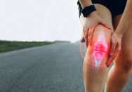 Eine Person hält sich das schmerzende rechte Knie, das von einer digitalen roten Grafik hervorgehoben wird, was auf Gelenkschmerzen oder Arthrose hinweist. Die Person befindet sich auf einer leeren Straße in einer ländlichen Umgebung.