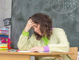 Eine Lehrerin sitzt an ihrem Pult vor der Tafel in einem Klassenzimmer. Sie fasst sich mit der rechten Hand an die Stirn und sieht erschöpft  aus.