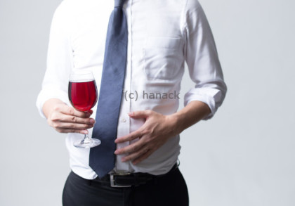 Ein Mann in einem weißen Hemd und einer blauen Krawatte hält ein Glas Rotwein in der Hand und berührt seinen Bauch mit der anderen Hand.