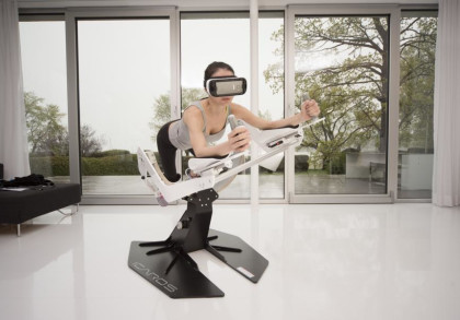 Eine Person steht in einem hellen Raum auf einem Trainingsgerät und trägt eine VR-Brille. Sie hält sich an den Griffen des Geräts fest, während sie sich nach vorne lehnt. Im Hintergrund sind große Fenster mit Blick auf eine Terrasse und Bäume zu sehen.