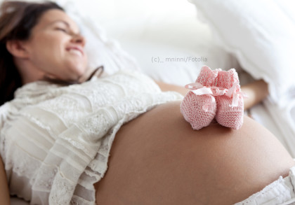 Ein Foto einer lächelnden schwangeren Frau in weißer Spitze, liegend auf einem Bett. Zwei kleine rosafarbene Babyschühchen liegen auf ihrem entblößten Babybauch.