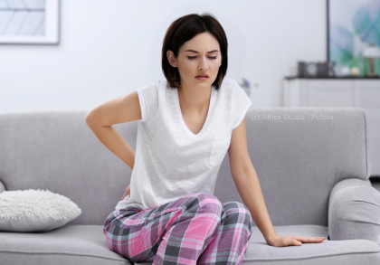 Eine Frau sitzt auf einem grauen Sofa, trägt ein weißes T-Shirt und karierte rosa-lila Pyjamahosen. Sie hält sich den unteren Rücken und zeigt ein schmerzverzerrtes Gesicht. Im Hintergrund befinden sich unscharfe Möbel und Dekorationen.
