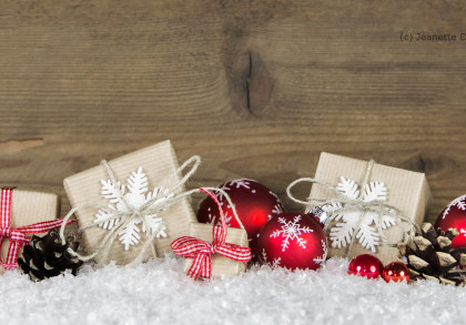 Geschenkboxen in braunem Papier verpackt und mit weißen Schneeflockendekorationen, roten Bändern und Juteschnur verziert, liegen auf einer Schneeschicht. Daneben befinden sich rote Weihnachtskugeln mit weißen Schneeflockenmustern und Tannenzapfen.