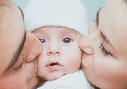 Ein Baby mit einer weißen Mütze wird von zwei Erwachsenen auf ihren Wangen geküsst.