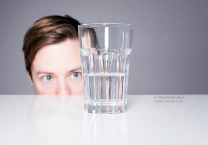 Ein Mann mit blauen Augen schaut neugierig über den Rand eines weißen Tisches. Vor ihm steht ein Glas, das zur Hälfte mit Wasser gefüllt ist. Der Hintergrund ist grau.