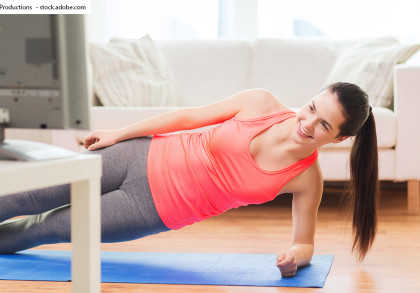 Eine Frau macht eine Seitplanke-Übung auf einer Yogamatte zu Hause vor einem Fernseher. Sie trägt ein neonorangfarbenes Tanktop und graue Leggings. Im Hintergrund sieht man ein Sofa mit bunten Kissen und Fenster, die viel Licht hereinlassen.