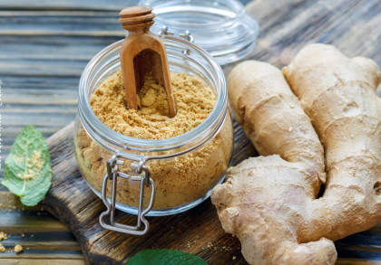 Ein Glas mit gemahlenem Ingwer, das auf einem Holzbrett steht. Neben dem Glas liegt frischer Ingwer und ein grünes Blatt. Eine kleine Holzschaufel steckt in dem Glas. Der Hintergrund besteht aus Holzbrettern.