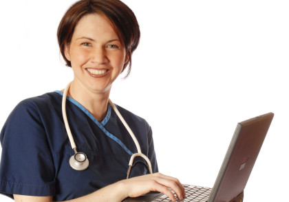 Eine lächelnde Frau in blauer medizinischer Arbeitskleidung, mit einem Stethoskop um den Hals, arbeitet an einem Laptop. Der Hintergrund ist weiß.
