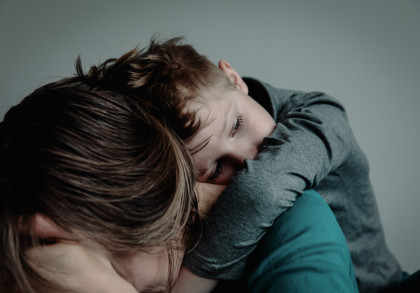 Ein kleiner Junge liegt mit geschlossenen Augen auf der Schulter einer erwachsenen Person mit braunen Haaren, die ihr Gesicht in den Händen vergräbt. Beide tragen graue Oberteile. Die Szene wirkt emotional und intim.