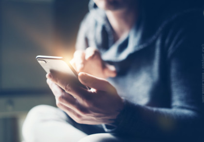 Eine Person in einem grauen Hoodie hält ein Smartphone in der linken Hand und tippt mit der rechten Hand darauf. Die Szene ist leicht unscharf, der Hintergrund wirkt verschwommen. Ein Lichtschein reflektiert auf dem Smartphone-Bildschirm.