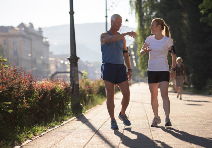 Zwei Personen gehen in sportlicher Kleidung auf einem sonnenbeschienenen Gehweg entlang. Sie scheinen sich zu unterhalten und beide tragen Armbänder für ihre Geräte. Im Hintergrund ist eine Stadtlandschaft mit Gebäuden und Grünflächen zu sehen.