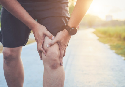 Eine Person, die sich an das Knie hält und Schmerzen hat, während sie auf einem Pfad im Freien steht. Die Person trägt eine Uhr und Shorts, und im Hintergrund ist ein sonniger Tag zu sehen.