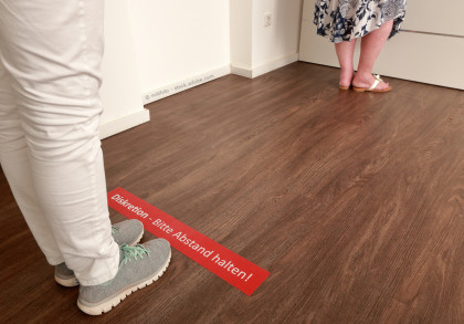 Zwei Personen stehen in einem Raum auf einem Holzboden. Die eine Person trägt weiße Hosen und graue Sneakers, die andere eine gemusterte Kleidung und weiße Sandalen.