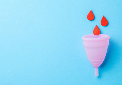 Das Bild zeigt eine rosa Menstruationstasse auf einem hellblauen Hintergrund. Über der Tasse schweben drei rote Tropfen.