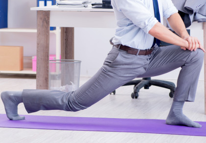 Ein Mann in Businesskleidung macht eine Dehnübung auf einer lila Yogamatte in einem Büro. Im Hintergrund sind ein Tisch mit Büromaterialien, ein Mülleimer und ein Regal zu sehen.
