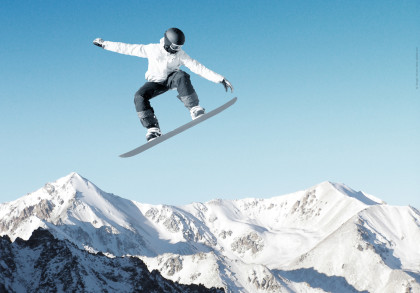 Ein Snowboarder in weißer Jacke und schwarzer Hose springt hoch über verschneite Berge bei klarem, blauem Himmel.