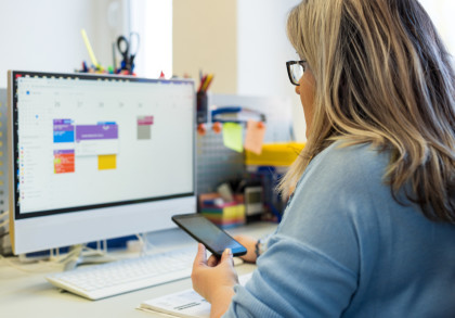 Eine Frau mit blonden Haaren und Brille sitzt an einem Schreibtisch und schaut auf einen großen Computerbildschirm, der einen digitalen Kalender anzeigt. In ihrer rechten Hand hält sie ein Smartphone.