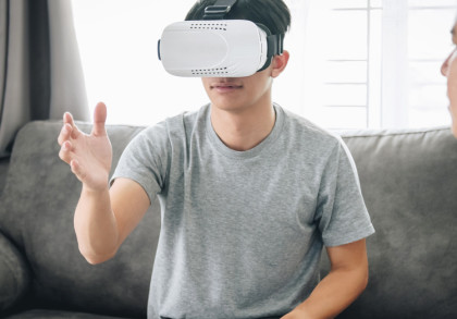 Ein Mann sitzt auf einem Sofa und trägt ein Virtual-Reality-Headset. Er hebt eine Hand, als ob er etwas in der virtuellen Welt berührt. Neben ihm sitzt eine weitere Person und beobachtet ihn. Im Hintergrund sind ein Fenster & ein grauer Vorhang zu sehen.