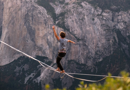 Eine Person balanciert auf einem Hochseil vor einer spektakulären Berglandschaft. Die Felswände im Hintergrund sind beeindruckend und steil.