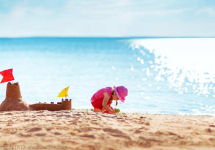 Zwei Kinder spielen am Strand. Das linke trägt ein gestreiftes Kleid und baut eine Sandburg mit einer roten Flagge. Das rechte trägt ein rotes Outfit & einen pinken Hut und spielt mit Sand. Der Hintergrund zeigt ein sonniges Meer mit glitzerndem Wasser.
