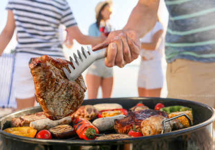 Nahaufnahme von einer Person, die ein BBQ-Werkzeug benutzt, um ein großes, gegrilltes Stück Fleisch von einem Grill zu heben. Auf dem Grill befinden sich verschiedene gegrillte Lebensmittel wie Gemüse, Würstchen und anderes Fleisch.