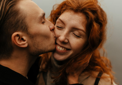 Ein Mann küsst eine lächelnde Frau auf die Wange. Die Frau hat rote Haare und Sommersprossen und ihre Augen sind geschlossen, während sie glücklich lächelt.