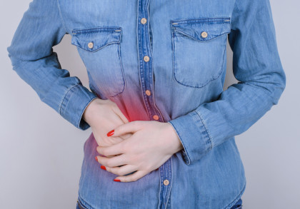 Eine Person in einem Jeanshemd hält sich den schmerzenden unteren Bauchbereich mit beiden Händen. Der Schmerzbereich ist durch eine rote Färbung hervorgehoben.