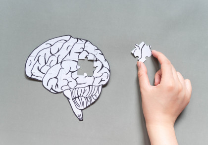 Das Bild zeigt eine Illustration eines Gehirns, das teilweise aus Puzzleteilen zusammengesetzt ist. Ein Puzzleteil fehlt, und eine Hand hält das fehlende Stück, um es in die Lücke zu setzen. Der Hintergrund ist grau.