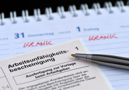 Eine Nahaufnahme zeigt eine Arbeitsunfähigkeitsbescheinigung und einen Kugelschreiber, die auf einem Kalender liegen. Im Kalender sind zwei Tage, Donnerstag und Freitag, mit der handschriftlichen Notiz „krank“ markiert.