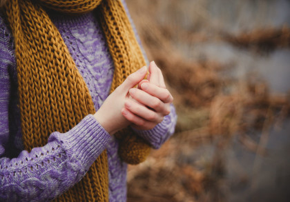 Nahaufnahme einer Person, die einen lila Strickpullover und einen senfgelben Strickschal trägt. Die Person hält ihre Hände vor dem Körper. Der Hintergrund ist unscharf und zeigt eine herbstliche Landschaft.