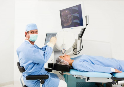 Dr. Breyer in blauer OP-Kleidung und Maske bedient eine Maschine während einer Augenoperation. Eine liegende Patientin trägt einen Schutzhaube, während ihr Auge behandelt wird. Ein Bildschirm zeigt medizinische Informationen.