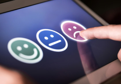 Eine Nahaufnahme eines Fingers, der auf einem Bildschirm ein trauriges Smiley-Symbol auswählt. Auf dem Bildschirm sind drei Smileys zu sehen: ein grünes lächelndes Gesicht, ein blaues neutrales Gesicht und ein lila trauriges Gesicht.