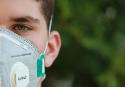 Nahaufnahme des Gesichts eines jungen Mannes mit grünen Augen, der eine graue Atemschutzmaske trägt. Der Hintergrund ist unscharf und zeigt grüne Farben.