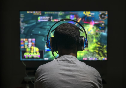 Ein Mann sitzt vor einem großen Bildschirm und spielt ein Computerspiel. Er trägt ein großes Headset mit blauen LED-Leuchten. Der Bildschirm zeigt eine bunte und detaillierte Spielszene mit vielen leuchtenden Farben.