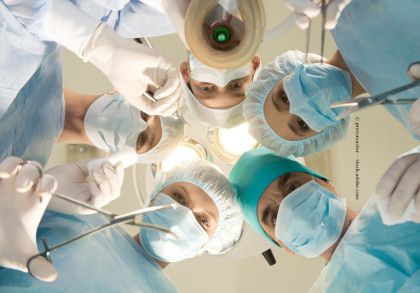 Das Bild zeigt eine Gruppe von Chirurgen in einem Operationssaal, die nach unten in die Kamera schauen. Sie tragen alle Operationskleidung inklusive Masken, Hauben und Handschuhen.