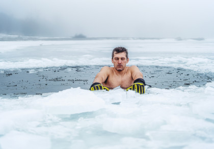Ein Mann ohne Hemd und mit gelben Handschuhen steht in einem gefrorenen See mit Eisstücken um sich herum. Der Mann hat die Augen geschlossen, und der Hintergrund ist neblig.