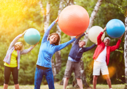 Eine Gruppe älterer Menschen macht Gymnastikübungen im Freien und hält dabei bunte Gymnastikbälle hoch. Alle sind in sportlicher Kleidung gekleidet und lächeln. Im Hintergrund sind Bäume und eine sonnige Atmosphäre zu sehen.