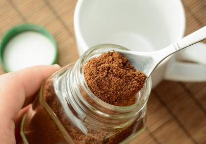 Nahaufnahme eines offenen Glases mit Instantkaffee, wobei ein Löffel mit Kaffeepulver über dem Glas gehalten wird. Im Hintergrund ist eine weiße Tasse und ein grüner Deckel zu sehen.