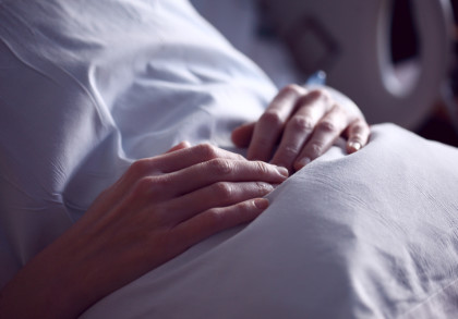 Nahaufnahme von zwei Händen, die auf einer weißen Bettdecke ruhen. Der Hintergrund ist unscharf und deutet auf eine Umgebung im Krankenhaus oder zu Hause hin. Die Hände wirken ruhig und entspannt.