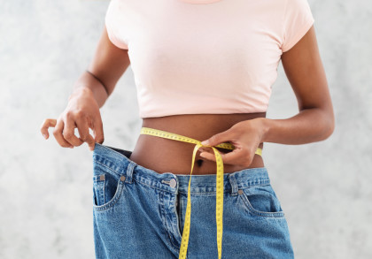 Eine Person trägt eine helle rosa T-shirt und hält ein Maßband um ihre Taille. Die Jeanshose, die sie trägt, ist locker und zeigt, dass sie Gewicht verloren hat.