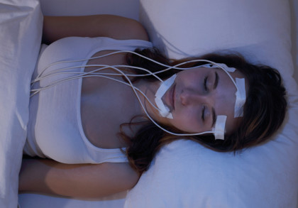 Eine Frau in einem weißen Tanktop liegt in einem Bett und schläft. Ihr Gesicht und Kopf sind mit medizinischen Elektroden und Kabeln verbunden, die mit Klebeband befestigt sind.