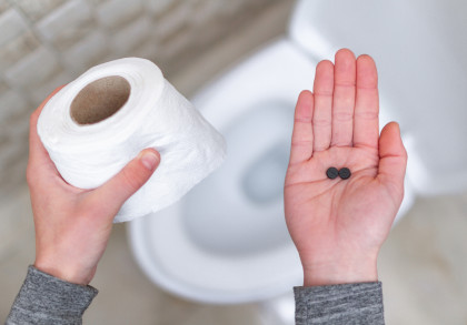 Nahaufnahme von zwei Händen: Die linke Hand hält eine Rolle Toilettenpapier, während die rechte Hand zwei schwarze Tabletten zeigt. Im Hintergrund ist eine Toilettenschüssel zu sehen.