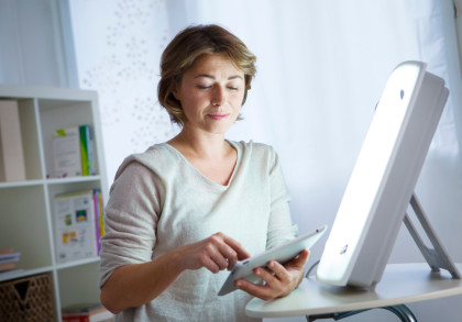 Eine Frau sitzt an einem Tisch und benutzt ein Tablet. Sie ist von einer hellen Lichtquelle beleuchtet, die möglicherweise eine Therapielampe ist. Im Hintergrund ist ein Bücherregal mit verschiedenen Gegenständen und Büchern zu sehen.
