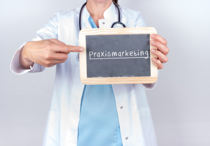 Ein Arzt oder eine Ärztin in einem weißen Kittel hält eine kleine Tafel mit der Aufschrift "Praxismarketing". Eine Hand zeigt auf die Tafel.
