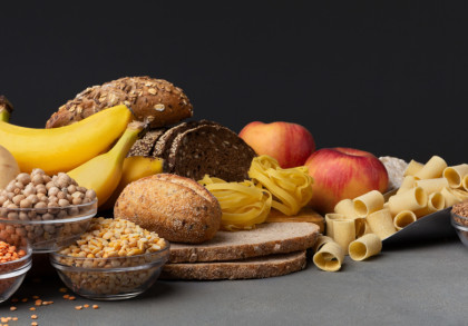 Eine Auswahl an kohlenhydratreichen Lebensmitteln auf einem Tisch vor einem dunklen Hintergrund. Zu sehen sind Bananen, Äpfel, Kartoffeln, verschiedene Arten von Brot und Brötchen, Nudeln sowie diverse Hülsenfrüchte und Getreide in Glasschalen.