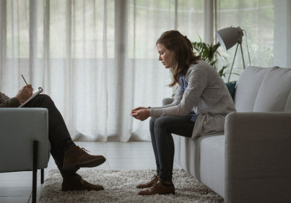 Eine Frau sitzt auf einem Sofa und beugt sich leicht nach vorne. Ihr Gesichtsausdruck wirkt nachdenklich und besorgt. Gegenüber von ihr sitzt eine Person, die ein Notizbuch hält und scheint Notizen zu machen.
