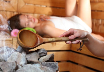 Ein entspannter Mensch liegt in einer Sauna. Eine Hand gießt Wasser aus einer Holzschöpfkelle auf heiße Steine, wodurch Dampf entsteht.