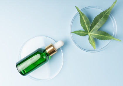 Ein grünes Glasfläschchen mit Pipette, liegend auf einem Petrischalen-Deckel, daneben eine große grüne Blatt-Pflanze in einer Petrischale, auf einem hellblauen Hintergrund.